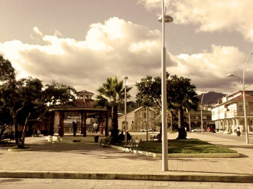 Stadtplatz mit Bäumen, Bänken und Menschen unter bewölktem Himmel.